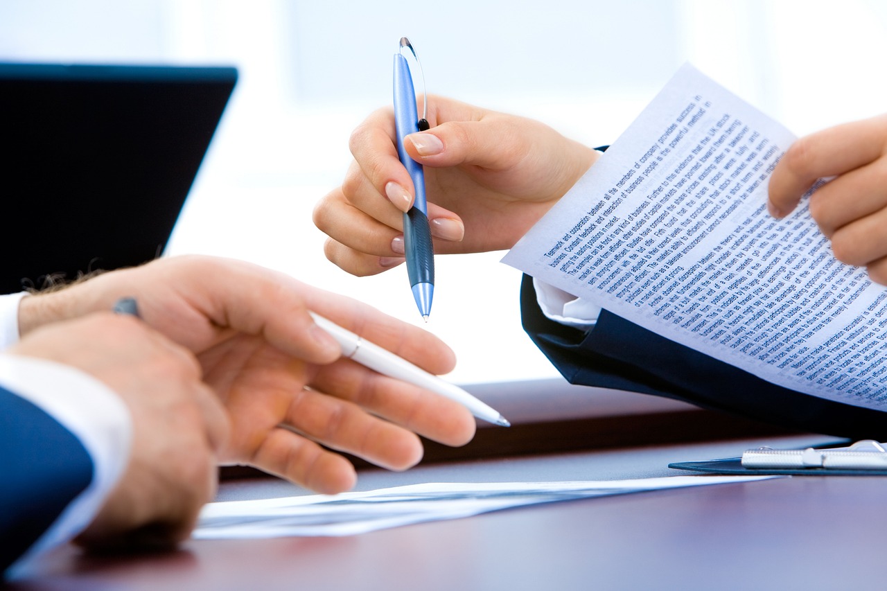 Mains autour d'un bureau, l'une tenant un stylo au-dessus d'une feuille imprimée, une autre feuilletant des documents