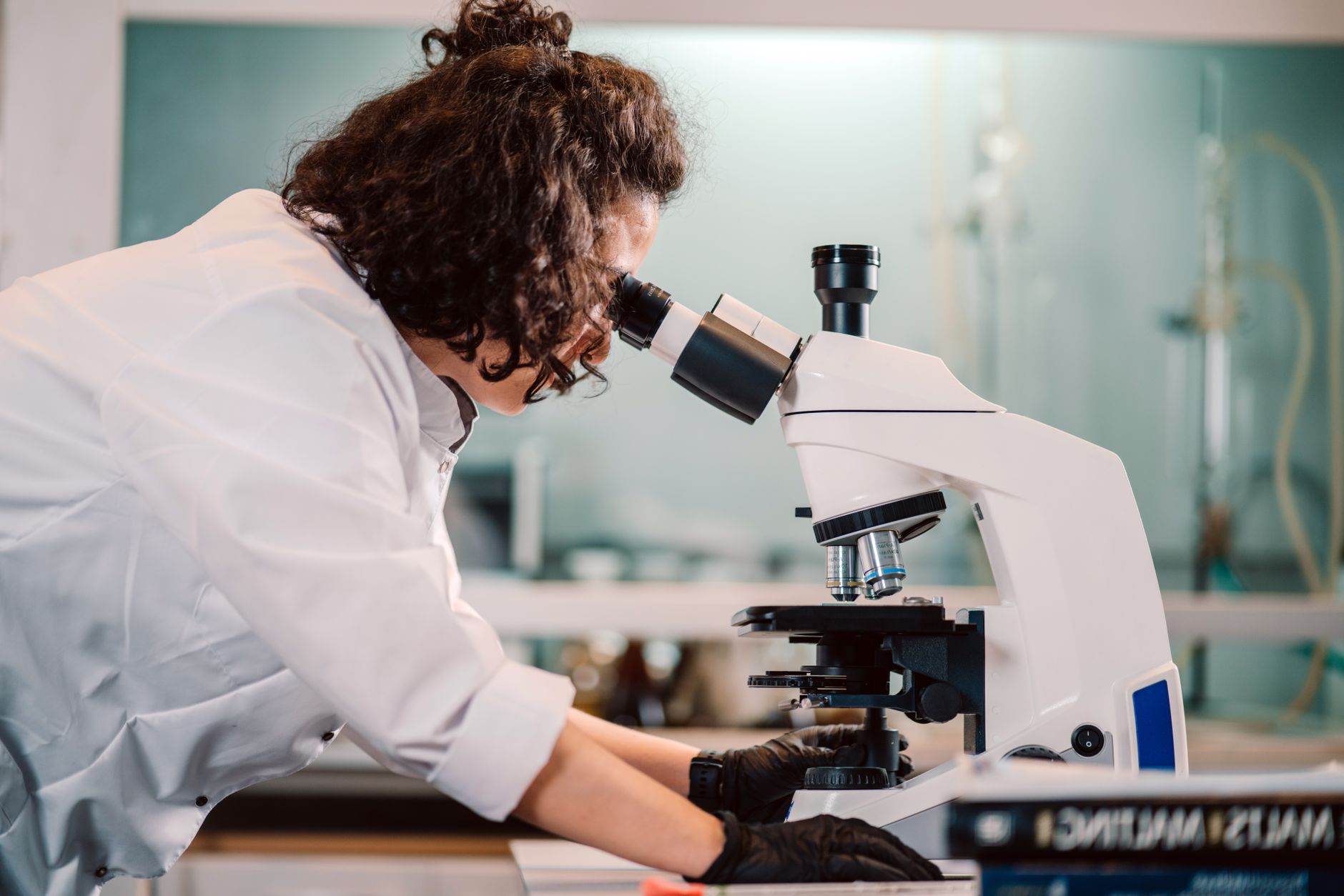 Une chercheuse en blouse blanche et gants noirs observe au microscope optique dans un laboratoire.