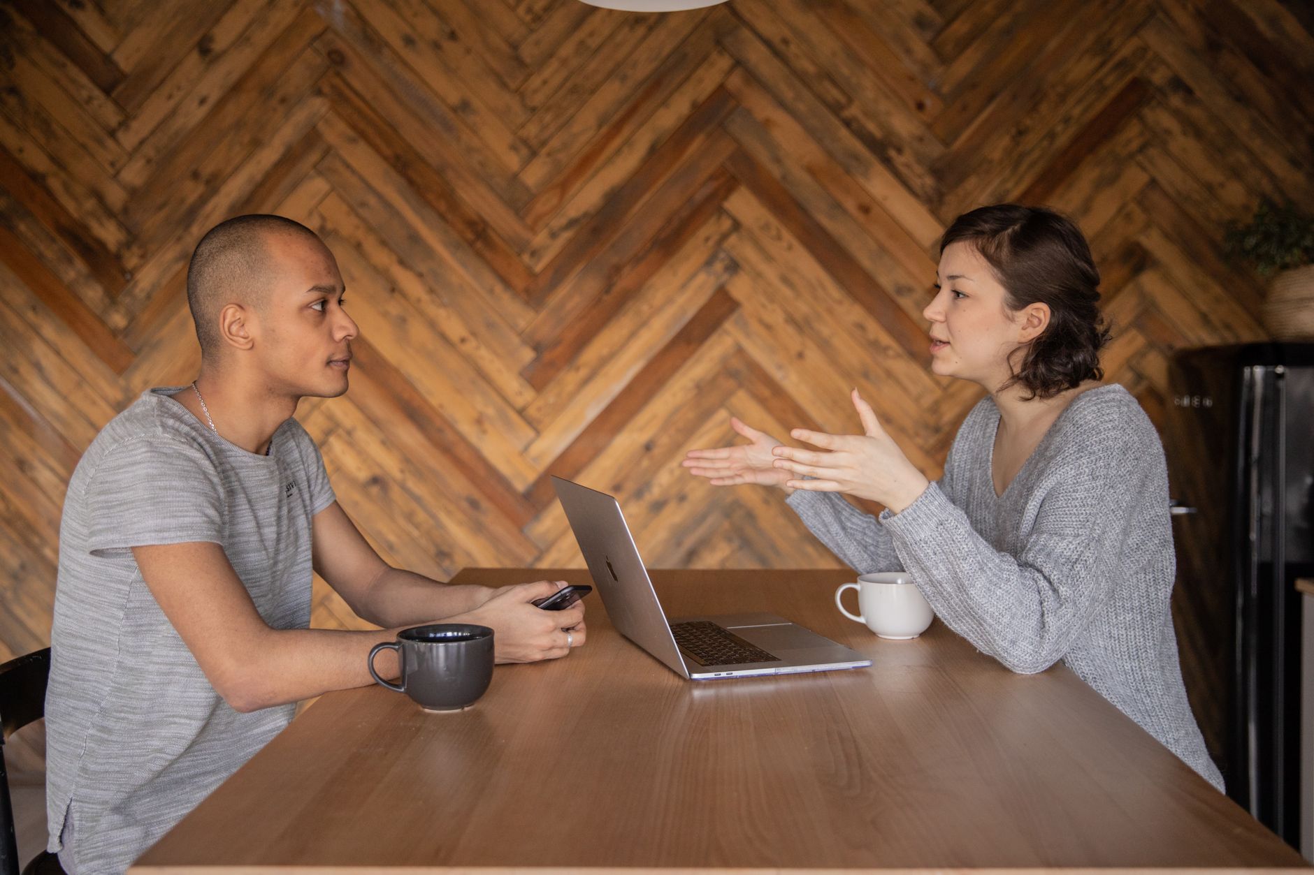 Une femme gesticule en parlant face à un homme qui l’écoute, de part et d’autre d’une table en bois avec ordinateur portable et tasses.